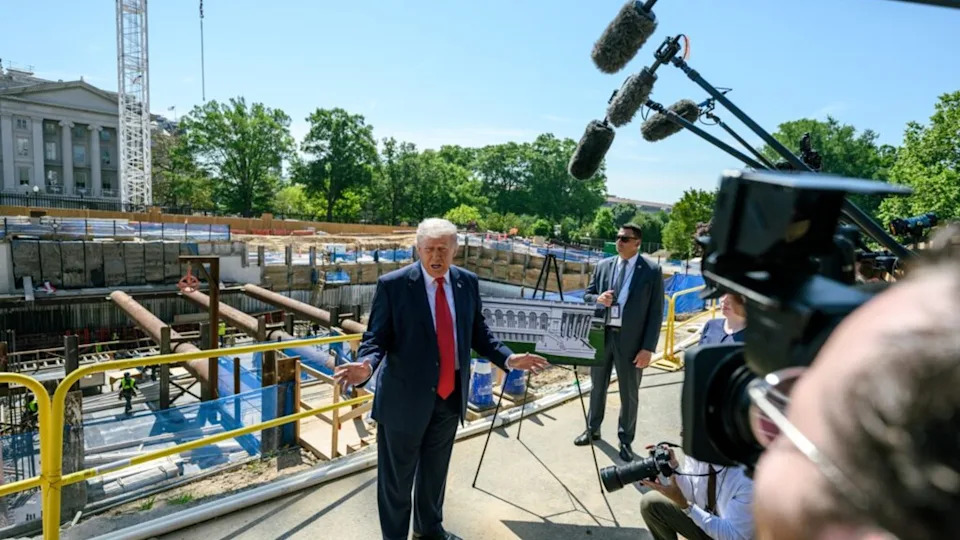 A person in a suit speaks to reporters at a construction site with a building in the background. (Credit: Image via Shutterstock)