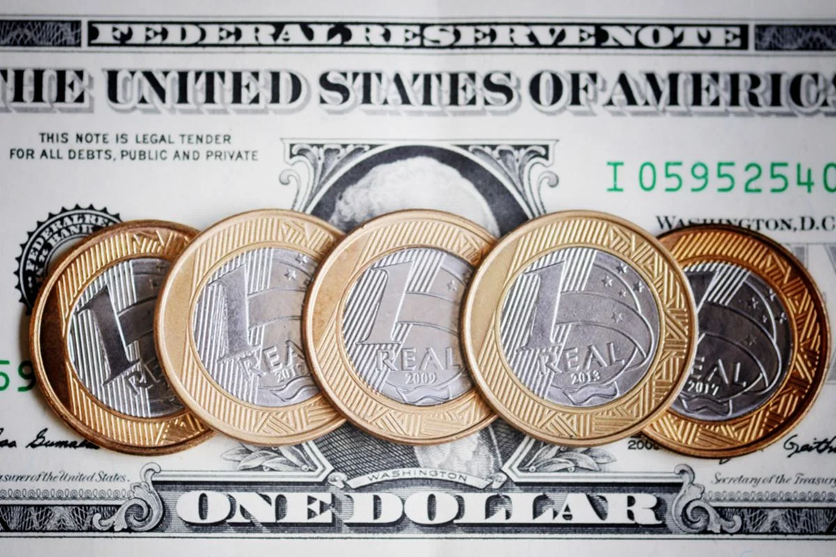 Imagem de moedas de real colocadas sobre uma nota de dólar - Metrópoles